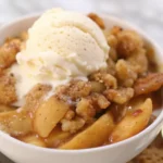 Warm apple cobbler topped with a scoop of vanilla ice cream in a white bowl.